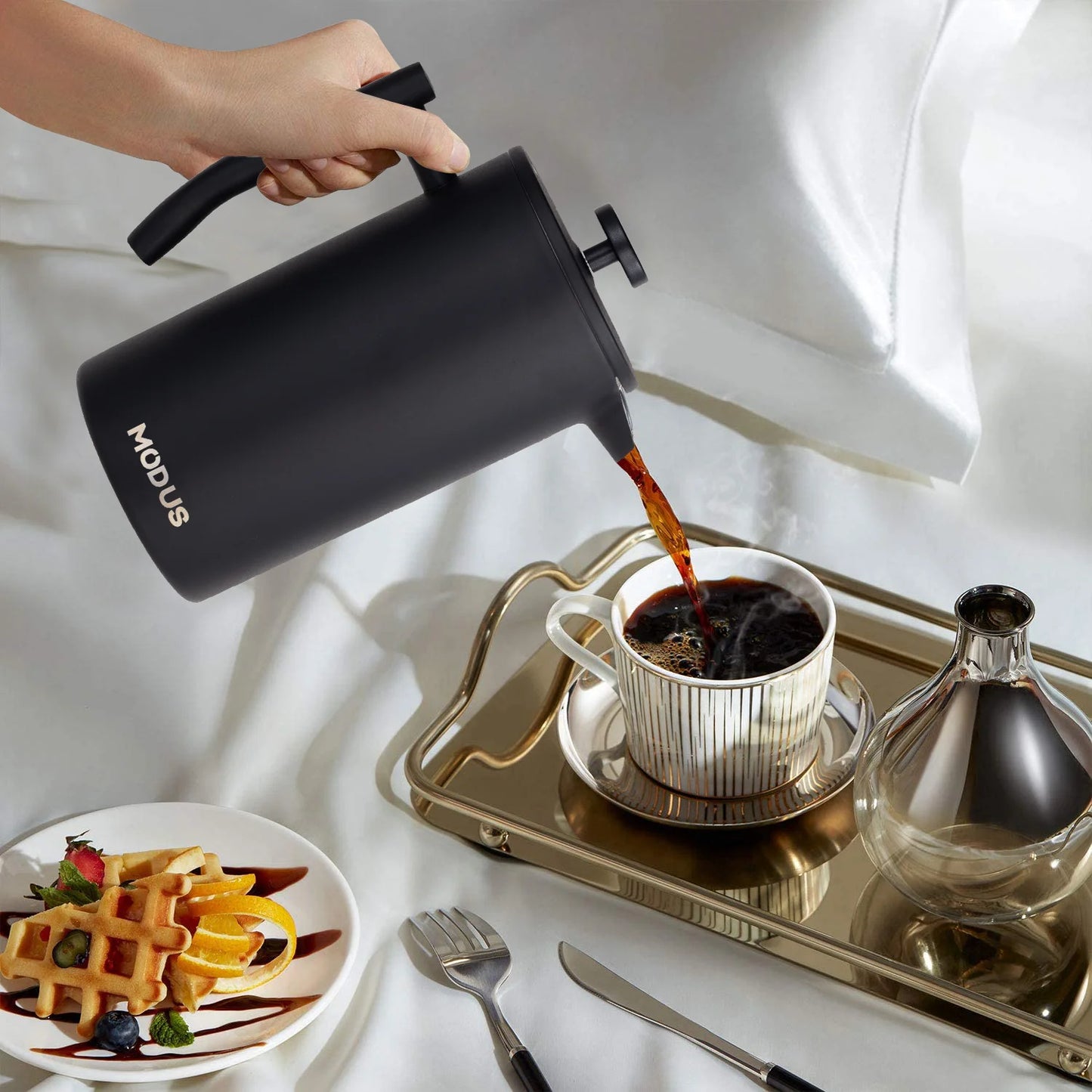 Cafeteira prensa francesa inox MODUS servindo café fresco em uma xícara elegante ao lado de um prato de waffles.