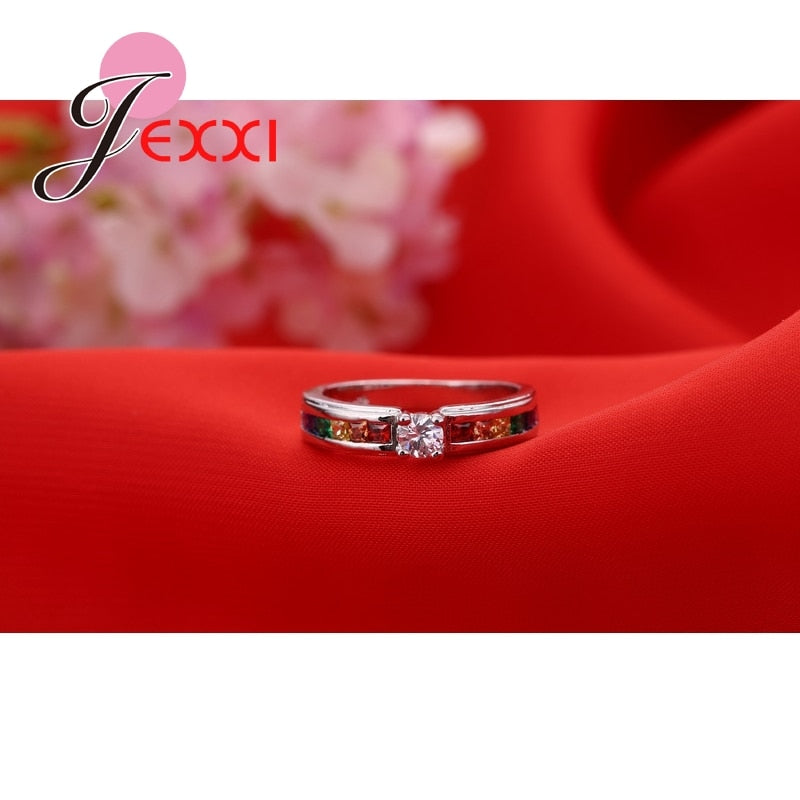 Anel de compromisso JEXXI com pedra central brilhante e detalhes coloridos em um fundo vermelho luxuoso