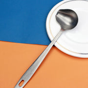 Colher concha escumadeira de inox LUCF com cabo ergonômico sobre prato branco e fundo colorido